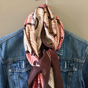 May include: A brown, pink, and white patterned silk scarf with a geometric design. The scarf is tied around a denim jacket.