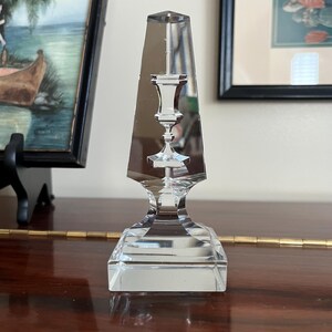 May include: A clear glass candlestick with a square base and a tall, pointed top. The candlestick has a decorative design in the center, resembling a smaller candlestick.