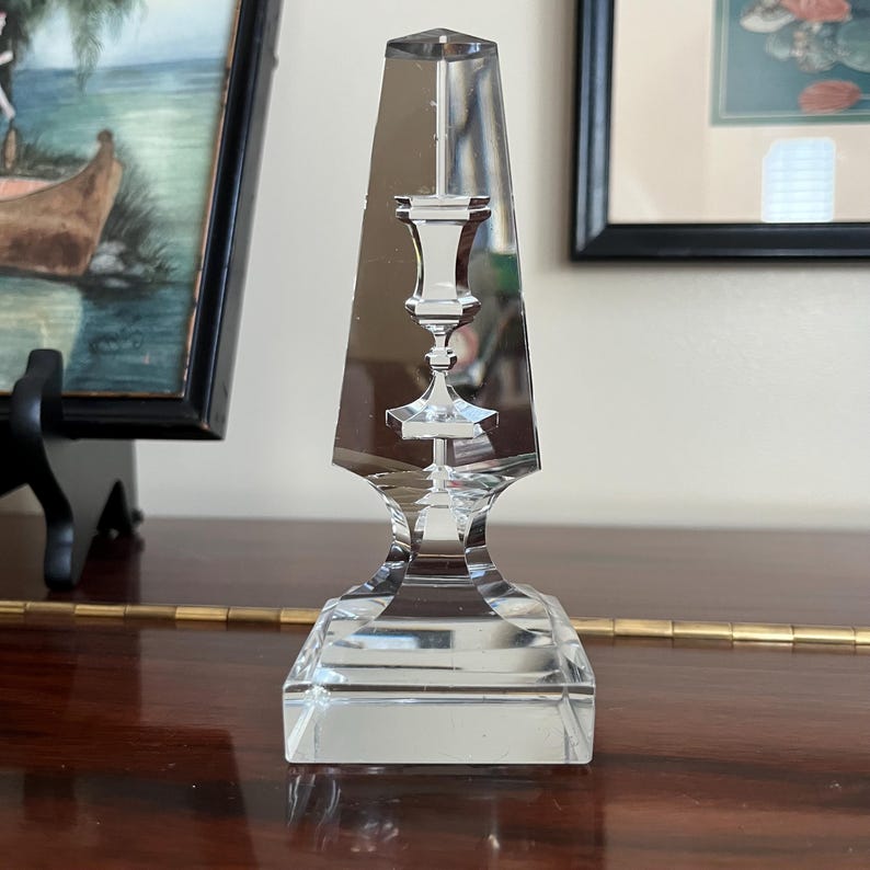 May include: A clear glass candlestick with a square base and a tall, pointed top. The candlestick has a decorative design in the center, resembling a smaller candlestick.