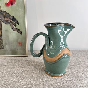 May include: A teal blue ceramic pitcher with a brown glaze detail and a unique, wavy design. The pitcher has a wide mouth and a handle.
