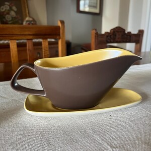 Puede incluir: Una salsera de cerámica marrón y amarilla con un platillo a juego. La salsera tiene un borde ancho y redondeado y una asa en el lado izquierdo. El platillo es de forma simple y redonda.