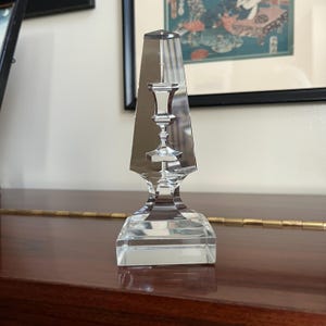May include: A clear glass candlestick with a geometric design. The candlestick has a square base and a tall, pointed top. The top of the candlestick is shaped like a vase.