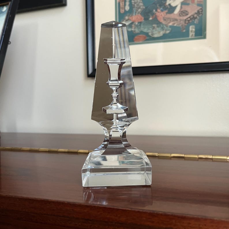 May include: A clear glass candlestick with a geometric design. The candlestick has a square base and a tall, pointed top. The top of the candlestick is shaped like a vase.