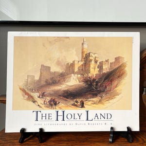 Puede incluir: Una impresión enmarcada de "The Holy Land" de David Roberts R.A. representa un paisaje detallado de una ciudad antigua con una torre alta. La impresión presenta una paleta de colores cálidos con figuras y camellos en una ladera. El título está impreso en una fuente serif clásica.