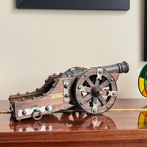 May include: A decorative wooden cannon with metal accents and wheels. The cannon has a dark brown finish with silver-coloured metal bands and rivets. The text "ARTILLERIA IMPERIAL CARLOS I" is visible on the side. The cannon is displayed on a polished wooden surface.