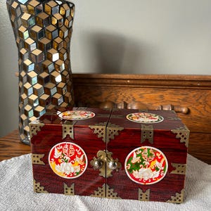 Può includere: Una piccola scatola di legno rossa con ferramenta in ottone e disegni circolari decorativi. La scatola ha un coperchio incernierato e una chiusura a fermaglio. Un vaso di vetro a mosaico è sullo sfondo.