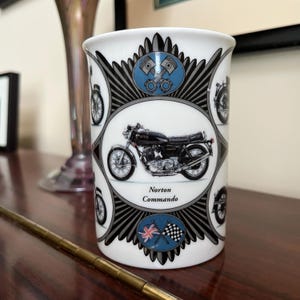 May include: White ceramic mug with a black and white design featuring a motorcycle and the text "Norton Commando".