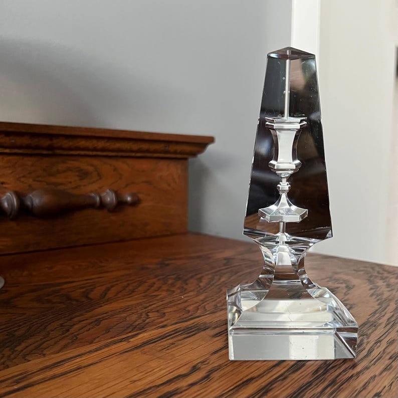 May include: A clear glass candlestick with a geometric design. The candlestick is shaped like a pyramid with a square base. The top of the candlestick is a small, round cup that would hold a candle.
