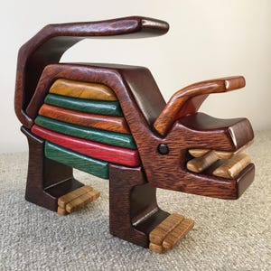 Puede incluir: Una escultura de animal de madera en forma de rinoceronte con una espalda rayada multicolor. La escultura está hecha de madera marrón oscura con un acabado brillante. El rinoceronte tiene un cuerno grande y una boca ancha con dientes.