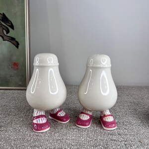 May include: Two white ceramic salt and pepper shakers shaped like people with pink shoes. The shakers have a pear-shaped body and are standing on two legs.