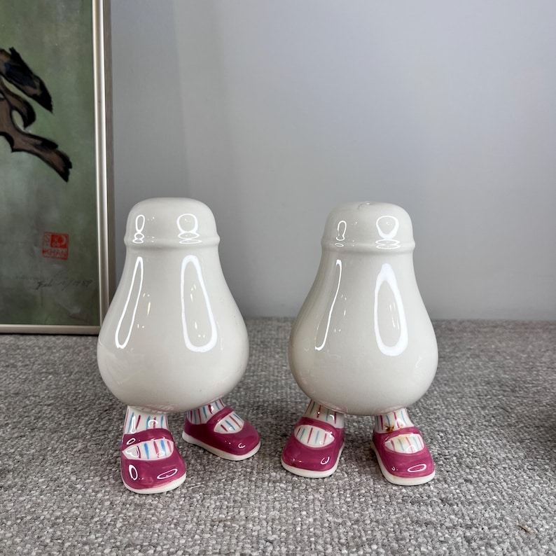 May include: Two white ceramic salt and pepper shakers shaped like people with pink shoes. The shakers have a pear-shaped body and are standing on two legs.