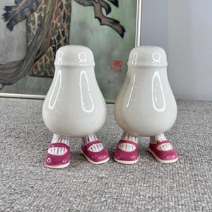May include: A pair of white ceramic salt and pepper shakers shaped like people with pink and white striped shoes.