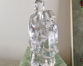 May include: A clear glass sculpture of two figures, possibly a couple, standing close together. The sculpture is abstract and has a modern design.