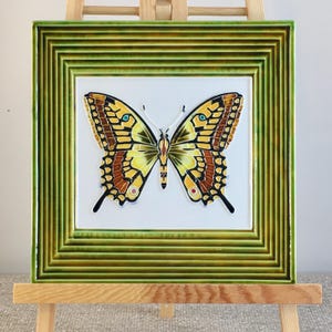 Può includere: Una piastrella di ceramica con una cornice verde e un'illustrazione di una farfalla gialla e nera.