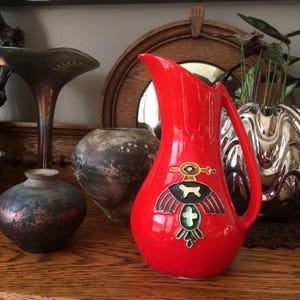 Peut inclure: Pichet en céramique rouge vif avec une anse et un emblème détaillé représentant un oiseau et une croix. Le pichet est rouge brillant et se distingue parmi d'autres vases décoratifs.