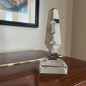 May include: A clear glass obelisk with a square base. The obelisk has a decorative design with a series of stacked circles.