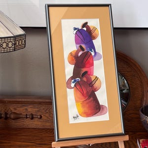 May include: A framed watercolor painting of four stacked pottery vessels. The vessels are painted in shades of purple, red, orange, and yellow. The painting is signed "Vera Rita" in the bottom right corner.