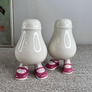 May include: A pair of white ceramic salt and pepper shakers shaped like people with pink shoes. The shakers have a glossy finish and are standing on a gray surface.