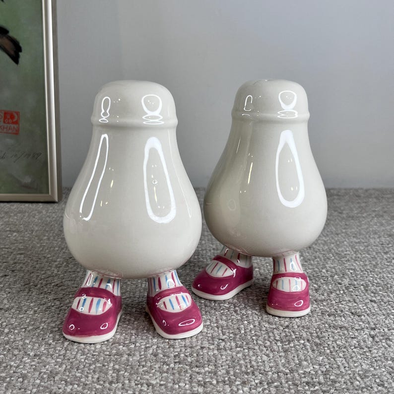 May include: A pair of white ceramic salt and pepper shakers shaped like people with pink shoes. The shakers have a glossy finish and are standing on a gray surface.