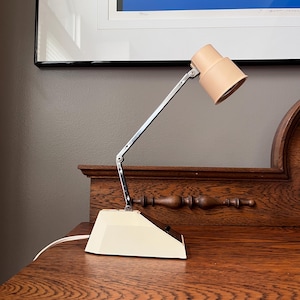 Może przedstawiać: Zabytkowa lampa biurkowa z kremową podstawą i beżowym kloszem. Lampa posiada chromowane ramię umożliwiające regulację. Lampa stoi na drewnianym biurku.