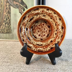 May include: A brown and white ceramic saucer with a textured, rippled surface. The saucer has a deep well in the centre and is displayed on a black stand.