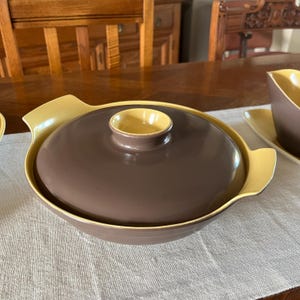 Puede incluir: Un plato de servir de cerámica marrón y amarillo con tapa. El plato tiene una forma redondeada y un borde ancho. La tapa tiene un pequeño pomo en la parte superior.