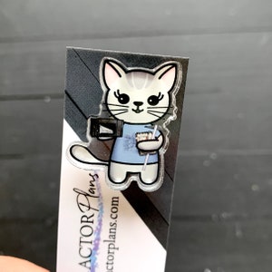 Pode incluir: Um marcador de página de acrílico transparente com um gato de desenho animado vestindo uma camisa azul e segurando uma xícara de café e um livro. O marcador de página tem o texto "ACTOR Plans" e "actorplans.com" impresso nele.
