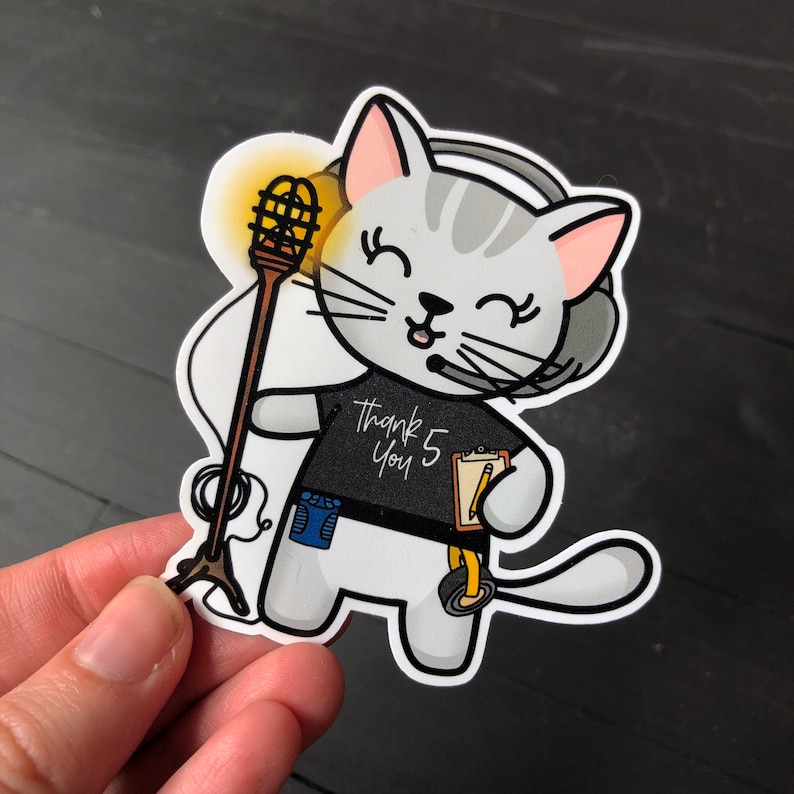 May include: A white cat with gray stripes wearing a black shirt with the text "Thank You 5" and a microphone headset. The cat is holding a clipboard with a pencil. The cat is holding a microphone with a yellow light on it.