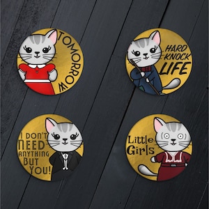 Può includere: Quattro bottoni rotondi dorati con gatti dei cartoni animati e testo. Il primo bottone dice "TOMORROW" e presenta un gatto con un vestito rosso. Il secondo bottone dice "HARD KNOCK LIFE" e presenta un gatto con un abito blu. Il terzo bottone dice "I DON'T NEED ANYTHING BUT YOU!" e presenta un gatto con un abito nero. Il quarto bottone dice "Little Girls" e presenta un gatto con un vestito rosso.