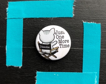 May include: White button with a cartoon cat sitting in a director's chair. The cat is wearing a name tag that says "Director". The text on the button reads "Just One More Time".
