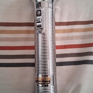 May include: A chrome-plated flashlight with a ribbed body and a polished head. The flashlight has a switch on the side and a label that reads "Sportsman". The flashlight is approximately 12 inches long.
