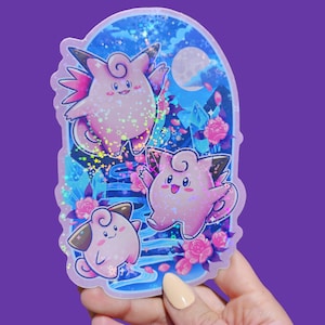 May include: A holographic sticker featuring three pink cartoon characters with wings, set against a blue and purple background with a full moon, stars, and pink flowers. The sticker has a glossy finish.