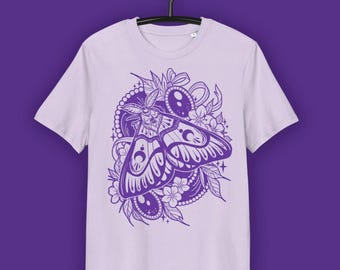 May include: A light purple t-shirt featuring a detailed purple moth design with floral and celestial elements. The moth has intricate wing patterns and is surrounded by flowers and decorative accents. The shirt is displayed on a hanger against a purple background.