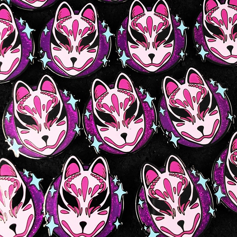 May include: Multiple pink and black enamel pins with a fox mask design on a purple glitter background. Each pin has a white outline and blue glitter stars.