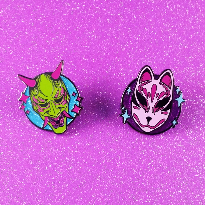 May include: Two enamel pins featuring Japanese masks. The first pin is green and features a demon mask with horns. The second pin is pink and features a fox mask with a swirl pattern. Both pins have a blue background with white stars.