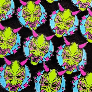 May include: A collection of green and pink enamel pins featuring a Japanese demon mask design. The masks have a detailed design with multiple eyes and a large mouth. The pins are set against a blue background with pink stars.