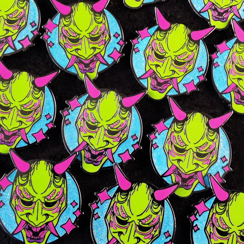 May include: A collection of green and pink enamel pins featuring a Japanese demon mask design. The masks have a detailed design with multiple eyes and a large mouth. The pins are set against a blue background with pink stars.