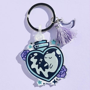 May include: A keychain featuring a heart-shaped bottle with a cat illustration inside. The bottle is surrounded by stars and flowers, with a purple tassel and a silver keyring. The design incorporates shades of blue, purple, and white.