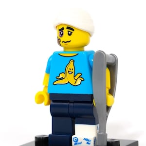 Peut inclure: Une figurine Lego représentant une personne avec un bandage blanc sur la tête, un œil au beurre noir et un pansement sur la joue. La figurine porte une chemise bleue avec un motif de banane jaune, un pantalon bleu et un plâtre blanc sur la jambe droite. La figurine tient une béquille grise. La figurine est debout sur une base noire.