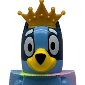 May include: A blue dog-like plastic toy figure with a yellow snout, large white eyes, and a gold crown on its head. The figure has a light blue belly patch, light blue arms and legs, and a pastel-colored cape attached around its neck area.