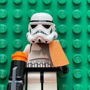 May include: A white LEGO Star Wars Stormtrooper minifigure with a black and orange blaster rifle and an orange cape. The minifigure is standing on a green LEGO baseplate.