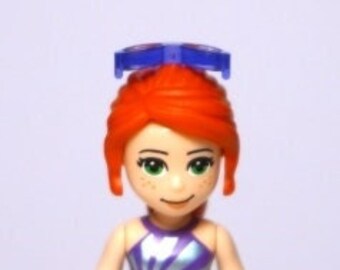 Puede incluir: Una minifigura Lego de una niña con el pelo rojo que lleva una camiseta morada y plateada, pantalones cortos de color verde lima y una diadema azul.