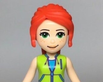 Puede incluir: Una minifigura Lego de una niña con el pelo rojo, que lleva un chaleco verde, pantalones cortos morados y zapatos azules.