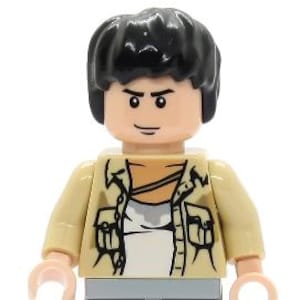 Pode incluir: Uma minifigura Lego com cabelo preto, uma jaqueta bege sobre uma camiseta branca e calças cinza claro. A figura tem uma cabeça cor de pêssego com um rosto impresso com sobrancelhas escuras e um sorriso, e braços e mãos de cor clara.