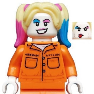 Op de afbeelding: Een Lego minifiguur van Harley Quinn die een oranje jumpsuit draagt met "ARKHAM ASYLUM" erop gedrukt. Ze heeft roze en blauwe staartjes en een ondeugende uitdrukking.