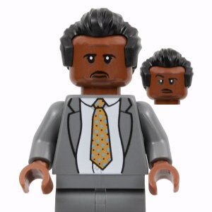 Puede incluir: Una minifigura Lego de traje gris con una pieza de pelo negro y una corbata con estampado amarillo y azul. La minifigura tiene un tono de piel marrón y una expresión seria.