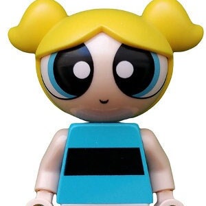 May include: A plastic toy figure of a girl with blonde pigtails, wearing a blue and black striped shirt, white pants, and black shoes. The figure has large blue eyes and a smiling face.