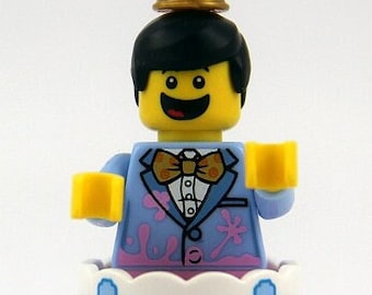 May include: A Lego minifigure wearing a blue tuxedo with pink splatter paint and a gold party hat, standing on a two-tiered cake with blue and white frosting.