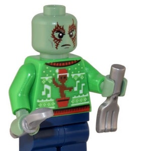 Puede incluir: Minifigura Lego verde con un suéter navideño verde y rojo, sosteniendo una herramienta plateada. La minifigura tiene una cabeza verde con marcas rojas y piernas azules. También se incluyen dos piezas Lego azules.