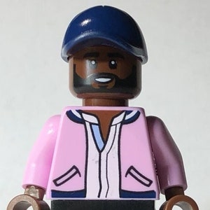 May include: A Lego minifigure with a dark brown skin tone, black hair, and a black beard. The minifigure is wearing a pink jacket with blue trim, a white shirt, black pants, and a dark blue baseball cap.
